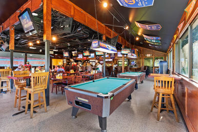 Lively sports bar interior with two pool tables, wooden high-top tables and chairs, multiple TVs and hanging team banners, patrons dining near the bar.