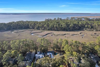Drone aerial view of a coastal marsh with a winding tidal creek, pine-lined waterfront homes in the foreground and a calm bay under a clear blue sky.
