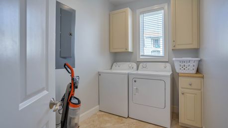 Bright residential laundry room with white top-load washer and front-load dryer under a window with blinds, beige wall cabinets, tiled floor, upright vacuum and a white laundry basket