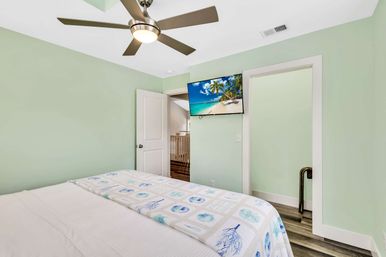 Bright mint-green bedroom with coastal quilt, modern ceiling fan, wall-mounted TV showing a beach, white trim and wood-look flooring, open door to hallway.