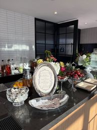 Stylish cocktail station on a black marble bar counter with a framed bar menu and silver trays, cocktail shaker, glass bowls of cherries and citrus garnishes, bottled spirits and fresh floral arrangements against white subway tile and dark paned doors.