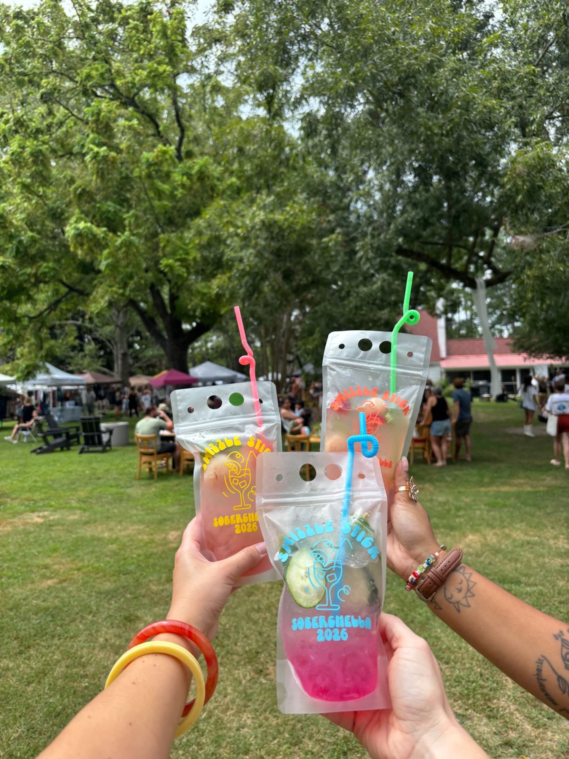 Three people cheers with colorful drink pouches and curly straws in a leafy park with vendor tents and festival crowds, outdoor summer market vibes.