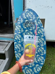 Hand holding a clear drink pouch with a pink curly straw in front of a blue floral skimboard covered in cream hibiscus prints, leaning against a trailer with metal steps and grass visible — tropical, beachy vibe.