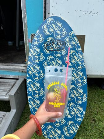 Hand holding a clear drink pouch with a pink curly straw in front of a blue floral skimboard covered in cream hibiscus prints, leaning against a trailer with metal steps and grass visible — tropical, beachy vibe.