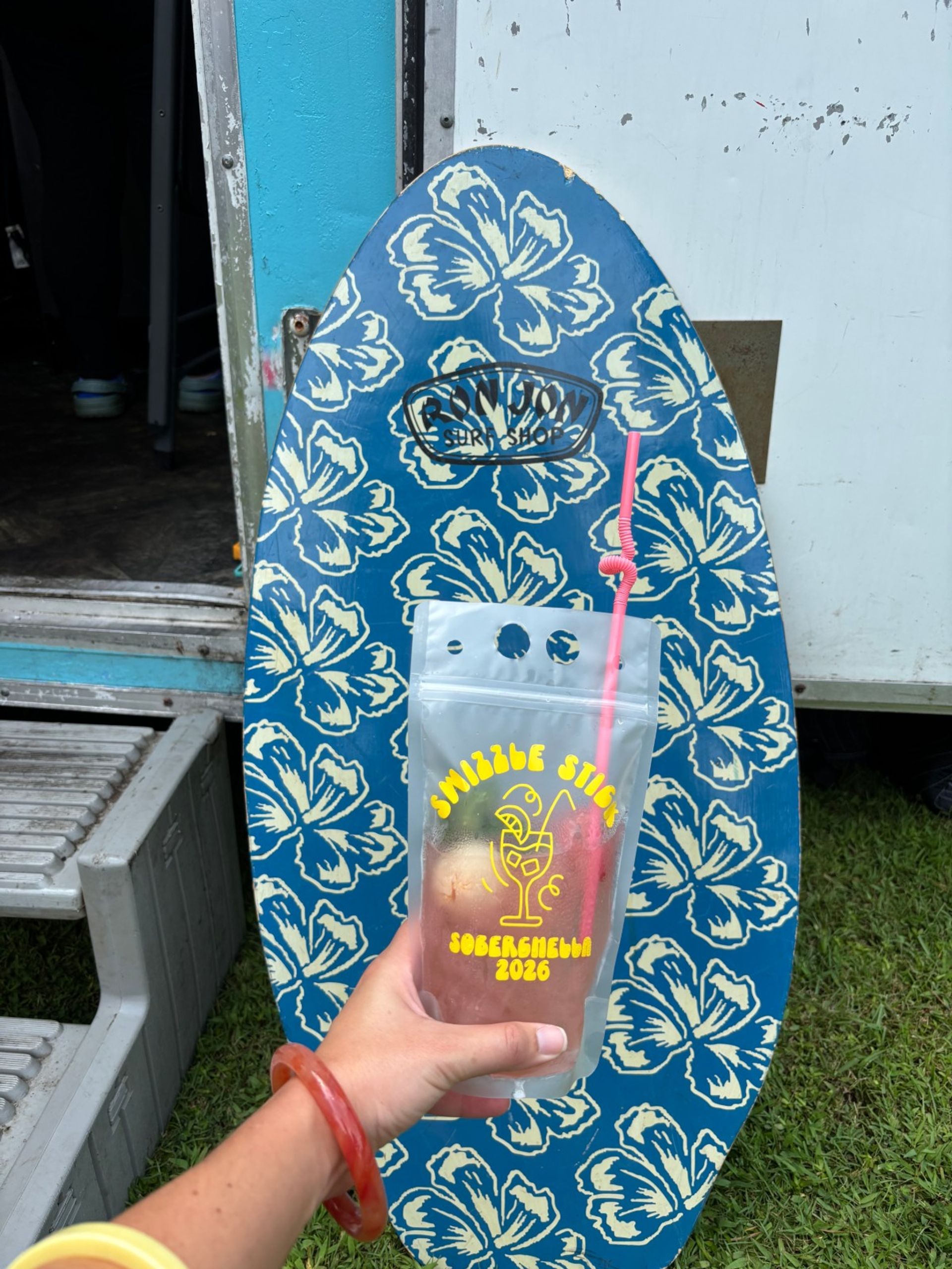 Hand holding a clear drink pouch with a pink curly straw in front of a blue floral skimboard covered in cream hibiscus prints, leaning against a trailer with metal steps and grass visible — tropical, beachy vibe.