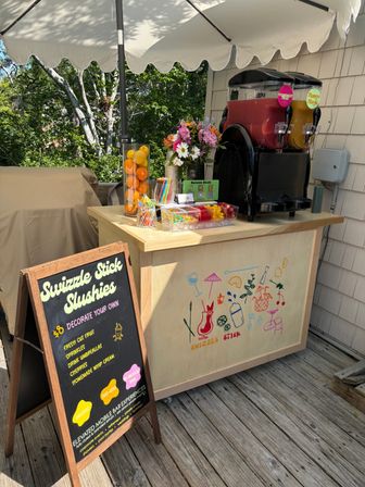 Sunny outdoor slushie cart on a wooden deck patio under a white umbrella, twin frozen drink dispensers (red and mango), jars of fresh citrus, colorful straws and toppings, chalkboard menu and flower bouquet.