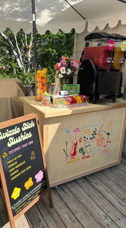 Sunny outdoor slushie cart on a wooden deck patio under a white umbrella, twin frozen drink dispensers (red and mango), jars of fresh citrus, colorful straws and toppings, chalkboard menu and flower bouquet.
