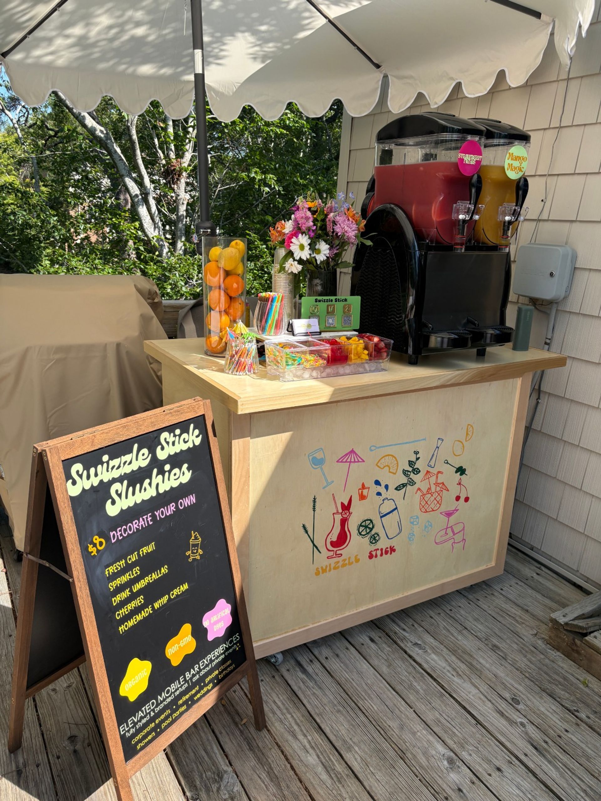 Sunny outdoor slushie cart on a wooden deck patio under a white umbrella, twin frozen drink dispensers (red and mango), jars of fresh citrus, colorful straws and toppings, chalkboard menu and flower bouquet.