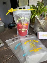 Clear resealable drink pouch with a yellow straw holding a red frozen mocktail, fun printed party graphic, fresh mint and tip jar on a wooden outdoor bar