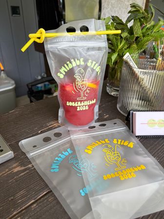 Clear resealable drink pouch with a yellow straw holding a red frozen mocktail, fun printed party graphic, fresh mint and tip jar on a wooden outdoor bar