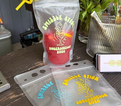 Clear resealable drink pouch with a yellow straw holding a red frozen mocktail, fun printed party graphic, fresh mint and tip jar on a wooden outdoor bar