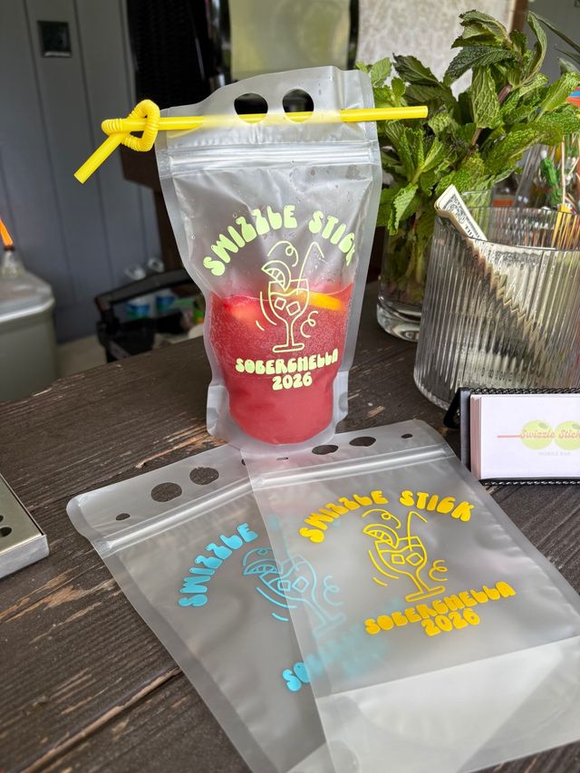 Clear resealable drink pouch with a yellow straw holding a red frozen mocktail, fun printed party graphic, fresh mint and tip jar on a wooden outdoor bar