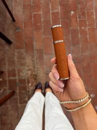 Stylish hand with French-manicured nails and gold bracelets holding a brown textured leather cylindrical case with silver trim over a terracotta brick patio, white pants and dark loafers visible below.