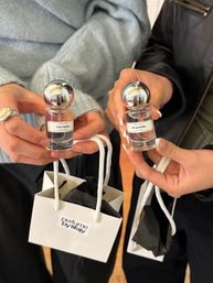 Close-up of two hands holding clear mini perfume bottles with reflective silver ball caps labeled “tea hehe” and “bb powda,” held above white shopping bags — boutique fragrance shopping lifestyle shot.