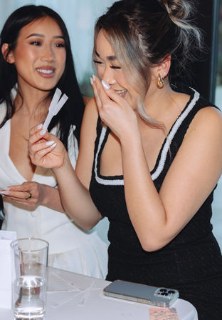 Two friends laughing at an indoor gathering, one holding perfume tester strips at a table with a smartphone and a glass of water.