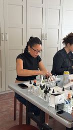 Participant concentrating at a fragrance workshop table in a bright studio, blending perfumes with glass dropper bottles, scent strips, notebooks and phones nearby.