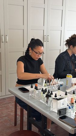 Participant concentrating at a fragrance workshop table in a bright studio, blending perfumes with glass dropper bottles, scent strips, notebooks and phones nearby.