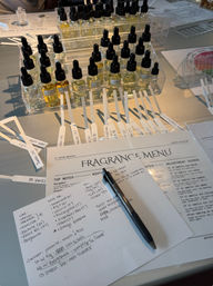 Perfume workshop table with rows of small dropper bottles, labeled scent strips fanned out, handwritten notes and a pen on a printed "Fragrance Menu" worksheet for scent testing