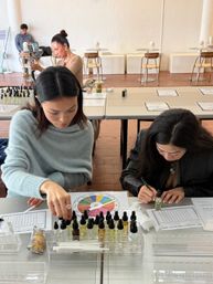 Two women at a bright studio table in a hands-on fragrance blending workshop, arranging dropper bottles, using a colorful scent wheel and recording notes.