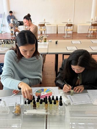 Two women at a bright studio table in a hands-on fragrance blending workshop, arranging dropper bottles, using a colorful scent wheel and recording notes.