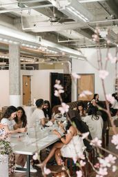 Bright urban loft studio with exposed ductwork and ceiling fans, pink blossom branches in the foreground, and a group at a long table taking part in a hands-on creative workshop with sample bottles and display stands.