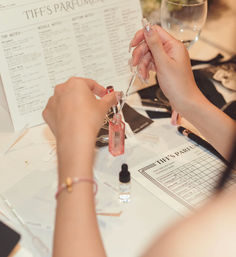 Hands with glittery nails using a pipette to fill a small pink perfume bottle at a DIY fragrance workshop, surrounded by scent notes charts, dropper vials and a tasting sheet on the table.