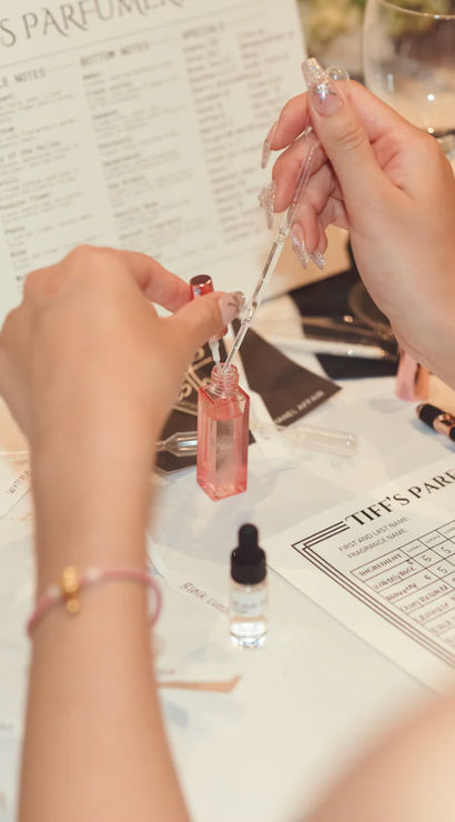 Hands with glittery nails using a pipette to fill a small pink perfume bottle at a DIY fragrance workshop, surrounded by scent notes charts, dropper vials and a tasting sheet on the table.