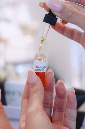 Manicured hands using a glass dropper to add golden frangipani oil into a small vial labeled "Frangipani Middle" in a spa-like beauty setting