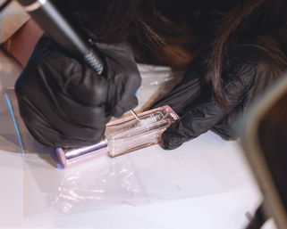 Close-up of black-gloved hands using a rotary engraving tool to etch a name into a small clear glass perfume bottle with a rose-gold cap, precision customization of a beauty accessory.