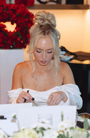 Elegant blonde in an off-shoulder white dress with a top‑knot bun, focused as she signs place cards at a flower‑lined reception table with a red rose display in the background.