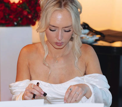 Elegant blonde in an off-shoulder white dress with a top‑knot bun, focused as she signs place cards at a flower‑lined reception table with a red rose display in the background.