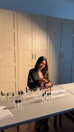 Sunlit studio scene of a woman at a worktable arranging small dropper perfume bottles and glass fragrance sample vials with beige paneled cabinets in the background.
