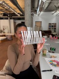 Smiling person holding a clear rack of perfume scent strips in a modern loft-style fragrance workshop, with notes, coffee and a scent wheel on the table.