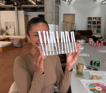 Smiling person holding a clear rack of perfume scent strips in a modern loft-style fragrance workshop, with notes, coffee and a scent wheel on the table.