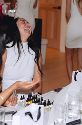 Laughing woman in a white sleeveless top at an indoor party, seated at a table filled with small dropper bottles, gift bags and glasses, with other guests in white dresses in the background.