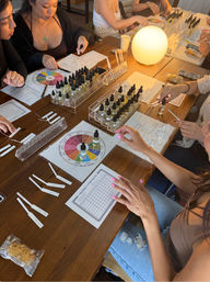 Perfume blending workshop at a wooden table — hands sampling scents from dozens of dropper bottles, scent wheel charts, fragrance blotters, worksheets and a warm glowing lamp in a cozy indoor class