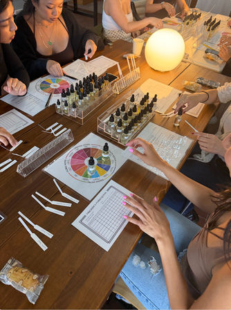 Perfume blending workshop at a wooden table — hands sampling scents from dozens of dropper bottles, scent wheel charts, fragrance blotters, worksheets and a warm glowing lamp in a cozy indoor class