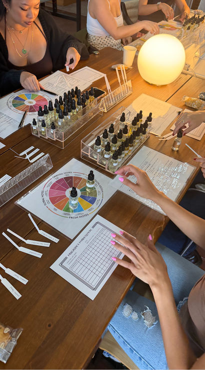 Perfume blending workshop at a wooden table — hands sampling scents from dozens of dropper bottles, scent wheel charts, fragrance blotters, worksheets and a warm glowing lamp in a cozy indoor class