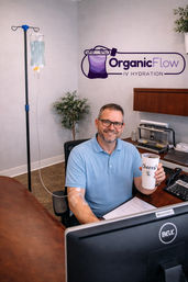 Smiling adult man seated in a calm IV hydration clinic receiving an IV infusion at his arm while working at a desk and holding a 'Cheers!' cup; visible IV bag on a pole, bandage on arm, computer and clinic logo on the wall.