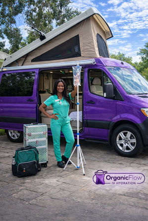 Mobile IV hydration nurse in teal scrubs poses with an IV pole and equipment cases beside a purple camper-style van with pop-up roof on a sunny driveway, showcasing mobile IV therapy services.
