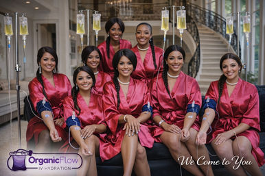 Eight smiling women in matching red satin robes sit on a sofa in a luxurious home interior with a sweeping staircase, each receiving IV hydration from hanging drip bags — glam, bridal-party style with braided hair and pearl necklaces.