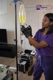 Clinician in purple scrubs wearing black gloves adjusts a yellow IV hydration bag on a stand during an at-home mobile IV therapy visit in a living room.