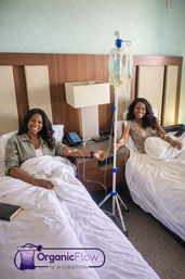 Two smiling women relax in adjacent beds receiving IV hydration drips from a shared pole in a bright, comfortable wellness room.
