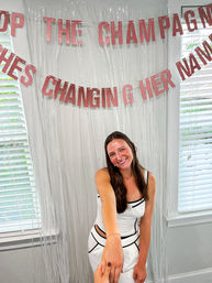Smiling woman in a white dress holding a hand at an indoor bachelorette/engagement party with pink glitter banner and window blinds backdrop