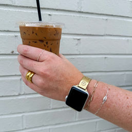 Close-up of a hand holding an iced coffee in a plastic cup with black straw against a white brick wall, featuring a gold smartwatch band, chunky gold ring and delicate silver bracelets.