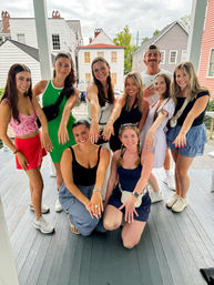 Group photo of nine friends on a wooden front porch by colorful houses, smiling and extending hands to show rings and manicured nails.