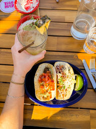 Sunlit outdoor patio scene: hand holding pineapple‑garnished margarita over two tacos with pico de gallo and slaw on a blue plate on a wooden table, lime wedge and water glasses nearby.