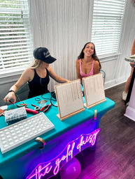 Two women at an indoor jewelry pop-up table with a teal cloth and pink neon script sign, necklace display boards, bead trays and tools; one woman smiling.