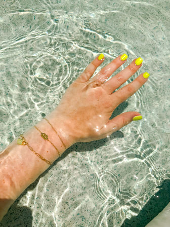 Close-up of a hand with neon yellow manicure and delicate gold bracelets in clear sunlit pool water, sparkling ripples and summer vibes