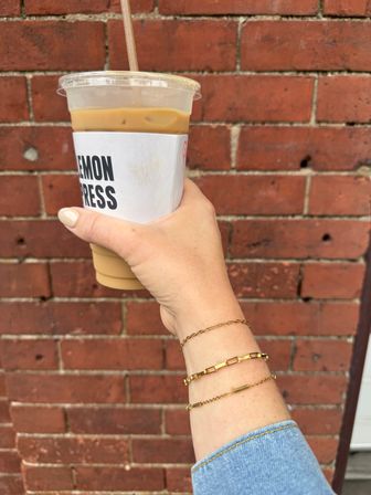 Hand holding iced coffee in a clear plastic cup with white sleeve, gold bracelets and denim cuff visible against an urban red brick wall